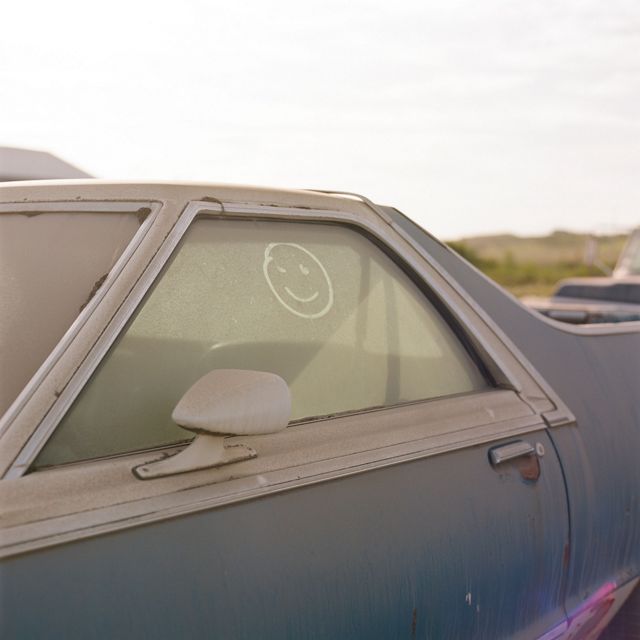 close up of a gray-blue older car covered in brown dust, a smiley face drawn in the window dust