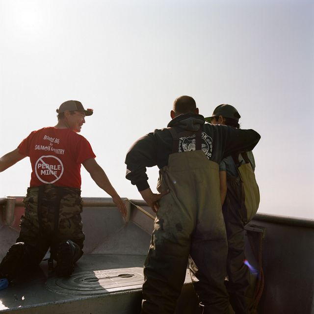 Two men and one boy stand in waders on the edge of a boat, backs turned, one with an arm around the boy.