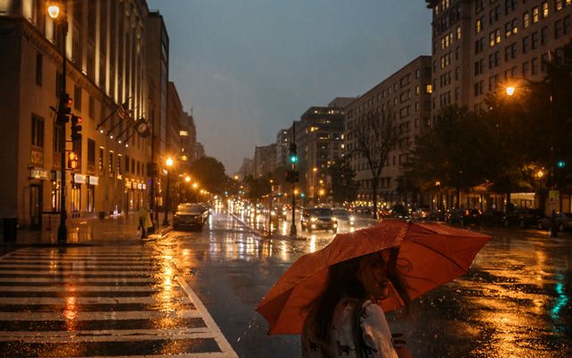 A woman holding a red umbrella waits to cross the street. Street lights and headlights are reflected on the wet pavement.