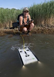 A woman crouches in the shallow water at the edge of a wetland. She holds a joystick pad in her hands, using it to deploy a small rectangular drone through the murky brown water.
