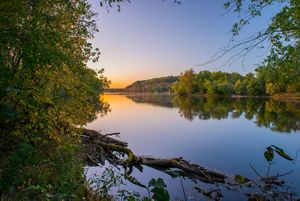 A sunsets over a river flowing between a forest.