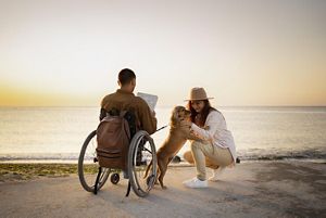 A family enjoying the beach during the sunset. There is a man in a wheelchair and a little dog playing with a woman.