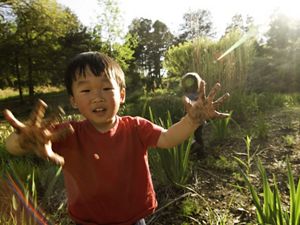smiling toddler stands in a green space with mud on his hands