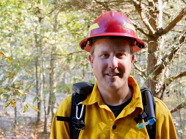 Gabe Cahalan headshot. A smiling man wearing yellow protective fire gear and a red hardhat stands in a forest during a controlled burn.