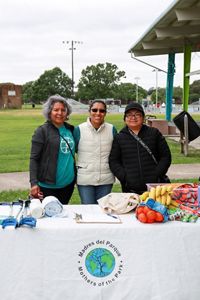 Three women stand together in a park behind a table with fruit.