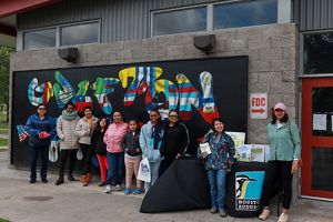 A group of men, women, and youths stand in front of a mural.