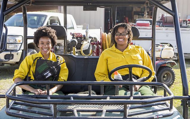 Two TNC Globe Interns dressed in yellow protective fire gear sit in a UTV in front of a fire engine, before a prescribed burn at Sandyland Sanctuary.