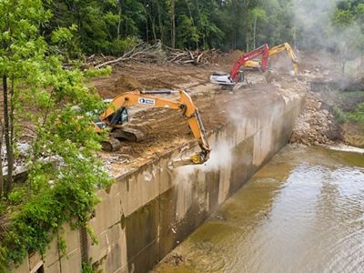 Large equipment hammers away at a large concrete structure on a river.