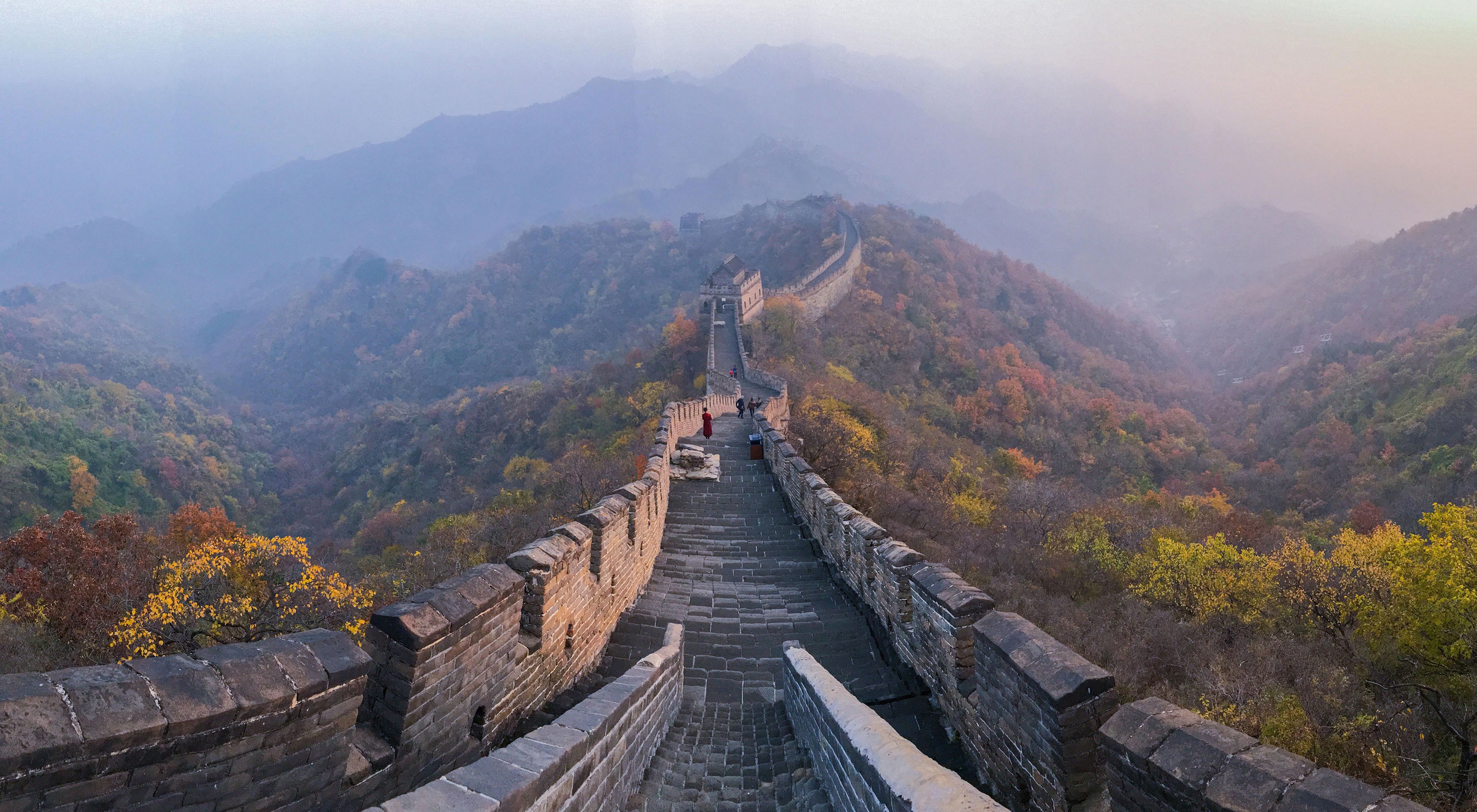 A view down the long pathway of the Great Wall of China shrouded in mist, with dense forest on either side of the wall.