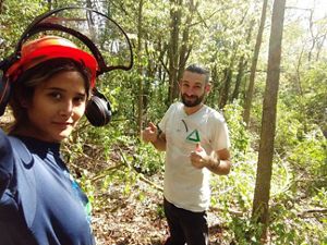 A woman wearing an orange helmet, standing in a forest, is looking at the camera, a man is standing beside her showing a thumbs up.