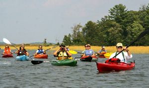 A group of adults in colorful kayaks are shown idling on a Maine lake, with binoculars and cameras.