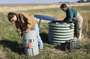 Staff members check drum-like water monitoring devices in the middle of a farmfield.