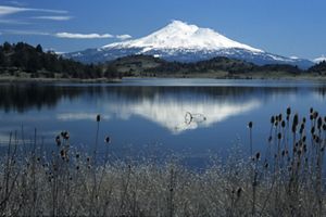 Snowcapped volcanic peak in the distance, reflected in the waters of a lake in the foreground.