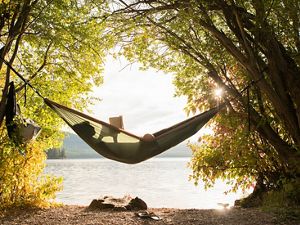 A woman reads a book while reclining in a hammock strung between two trees at the edge of a placid lake.