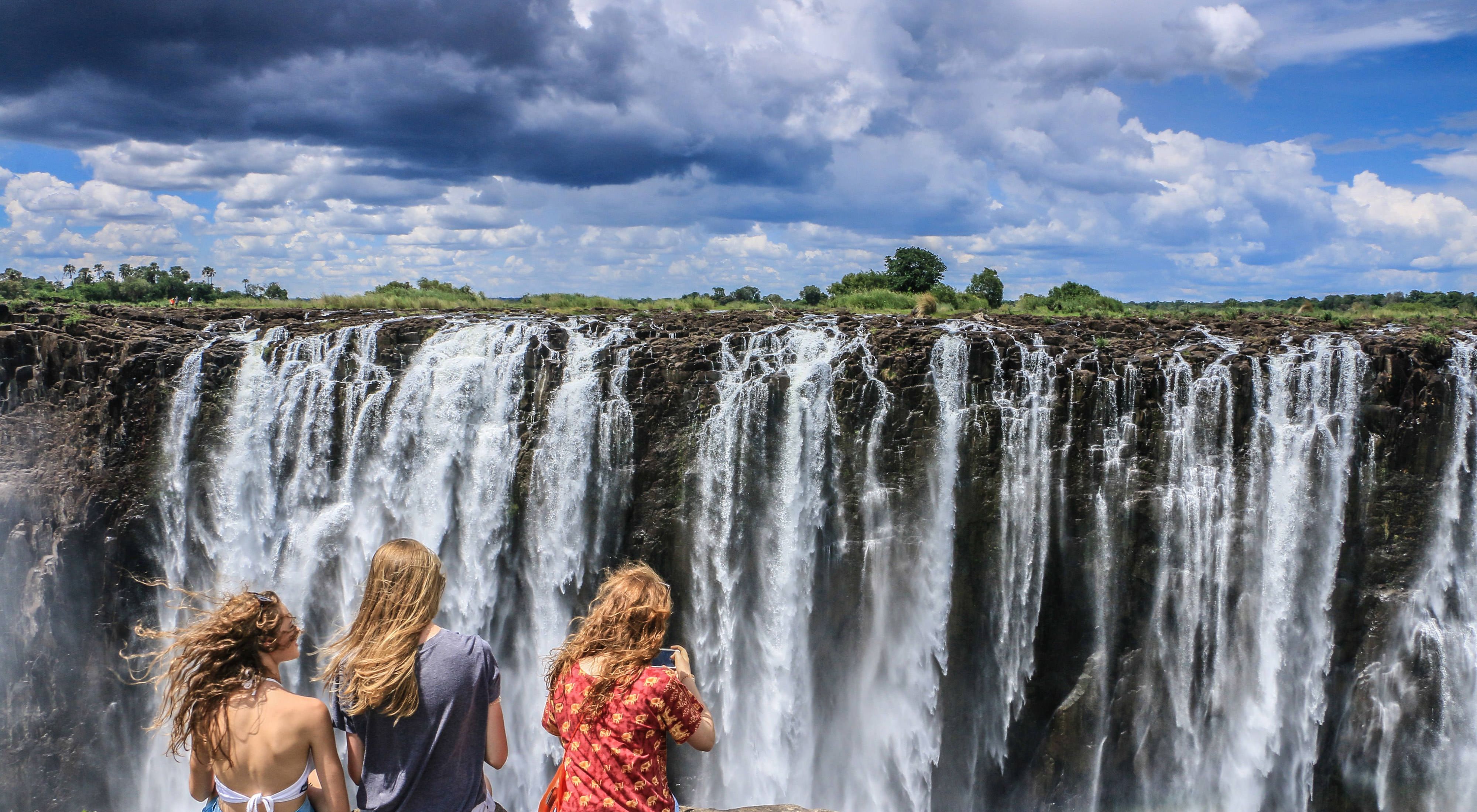 Three young people sit facing the Victoria Falls waterfalls as the wind blows their hair.