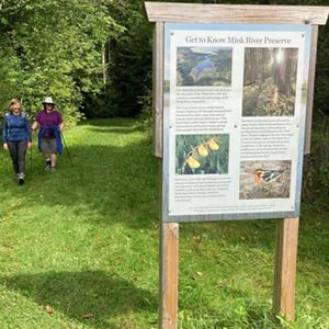 Two woman hike in the woods on a grassy path and approach a preserve sign that says 'Get to Know Mink River Preserve.'