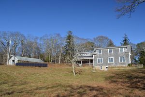 A gray shingled house sits on a grassy hill with a solar panel array to the left of it and trees and blue sky in the background.