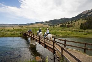 Five people on horseback cross a narrow wooden bridge over a stream, with green rolling hills and mountains in the background.