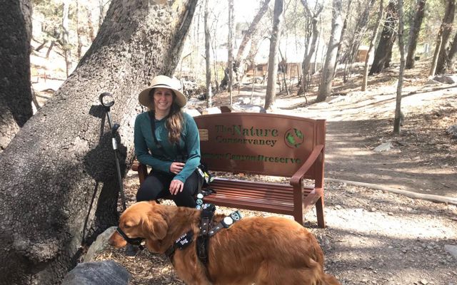 A woman is sitting on a TNC preserve bench with her service dog standing close by her. 