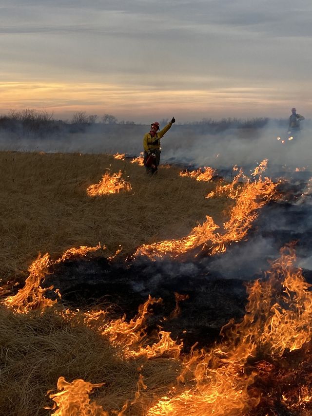 Fire crew member flashing the 'thumbs up' with burning grasslands in the foreground.