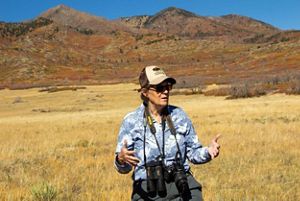 Terri Shulz stands in a grass field and presents to a group behind the camera, with mountains in the background.