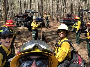 A woman holds up a camera to capture a group selfie of the six women standing behind her during a fire learning exchange. They are standing in a forest next to two ATVs.