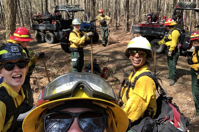 A woman holds up a camera to capture a group selfie of the six women standing behind her during a fire learning exchange. They are standing in a forest next to two ATVs.
