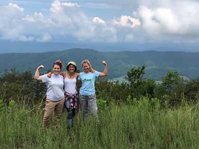 Three young woman stand shoulder to shoulder with their arms draped over each other. A tall green mountain ridge rises in the background behind them. Puffy white clouds hang in a blue sky.