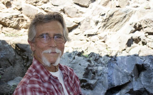 Portrait of a older man with graying hair and white beard. He is standing in front of a tall rock face.