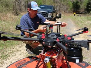 A man crouches on the ground next to a large drone, readying it for deployment on a controlled burn. The drone is sitting on a round orange landing pad.