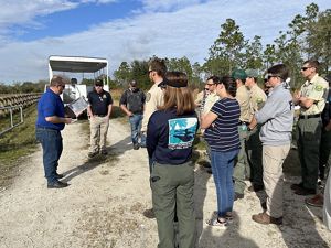 A group of people standing on a gravel road listening to someone speak.
