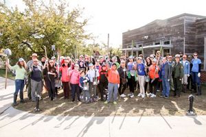 A large group of people standing outside of a building on the right hand side of the photo with green trees featured in background. Most of the people are holding gardening tools and smiling.