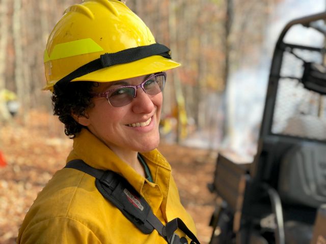 Deborah Landau headshot. A smiling woman wearing yellow protective fire gear and a yellow hard hat. White smoke rises behind her from a small fire.