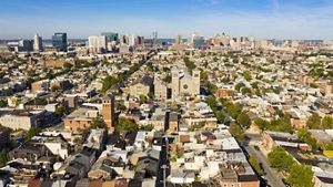 Aerial view of Baltimore looking towards the downtown skyline and Inner Harbor. The foreground is filled with rowhouse lined residential neighborhoods and centered on a large church.