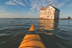 An abandoned farm house sits precariously at the edge of the water as the Chesapeake Bay slowly consumes it. The yellow nose of a kayak is visible in the foreground.