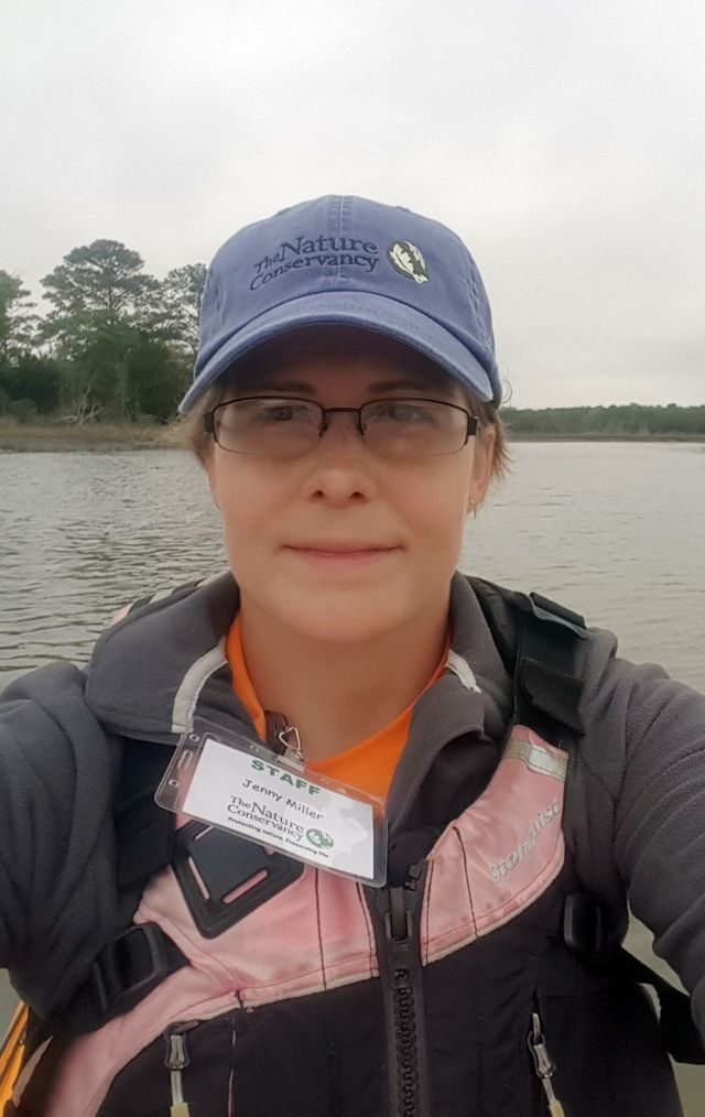 Selfie portrait taken by a woman sitting in a kayak. She is floating in a narrow coastal bay with wetlands and trees behind her.