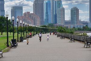 A city sidewalk stretches out next to a park lined with trees and green grass on either side. 