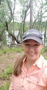 A selfie of Julia wearing a TNC baseball cap in front of a wetland