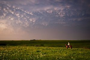 A man on horseback crosses a wide landscape of the Flint Hills of Kansas at dusk.