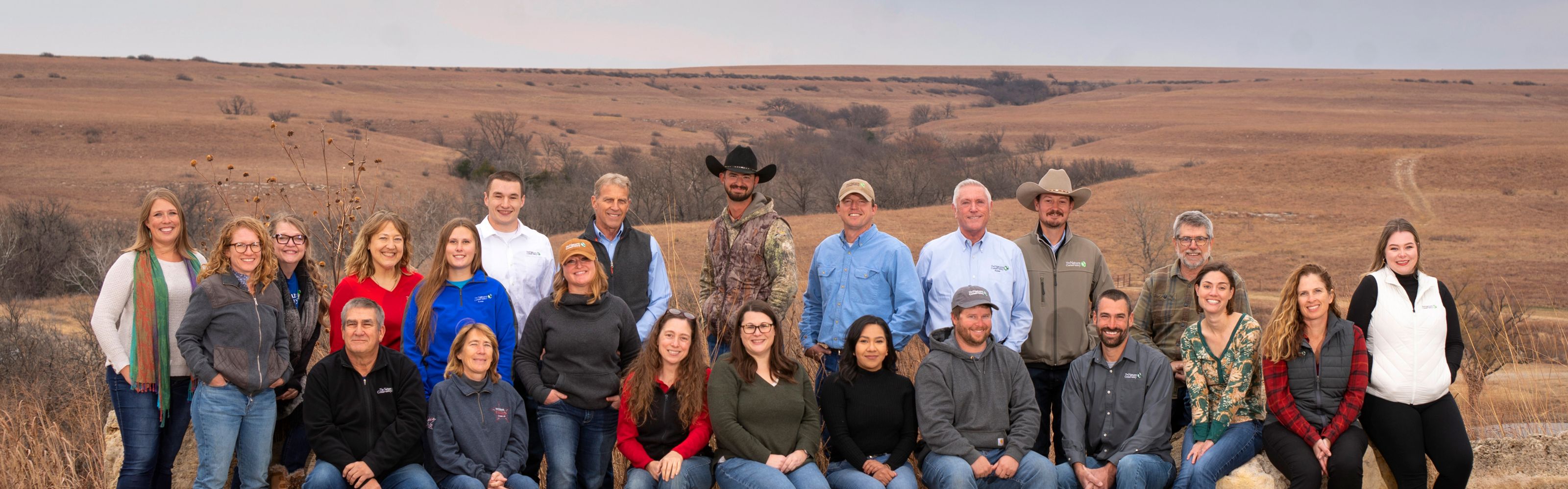 Group photo of about 25 staff members sitting and standing outside with prairie in the background.