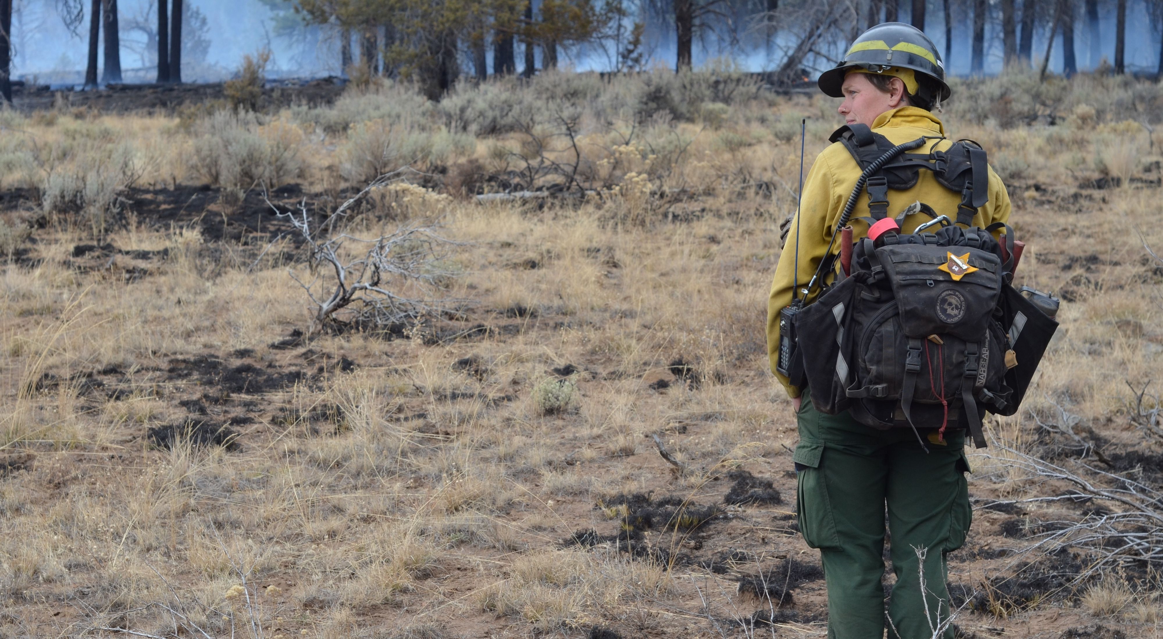TNC Oregon Burn Boss Katie Sauerbrey stands in fire practitioner gear with her back turned and a forest in front of her.