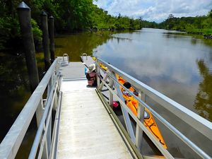 View from the end of a narrow floating dock with high metal sides. The dock leads to a path that disappears into a thick forest. Trees line the banks of the wide river that stretches to the horizon.
