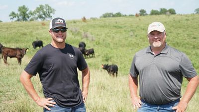 Two ranchers stand in a field with cows in background.