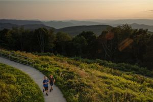 Two children run down a concrete path surrounded by grasslands and forest.
