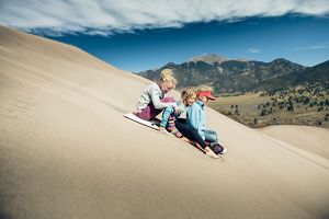 Three kids surf down sand dune. 