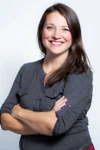 Kristin Bramell headshot. A smiling woman wearing a dark gray blouse poses in front of a white backdrop with her arms casually folded across her chest.