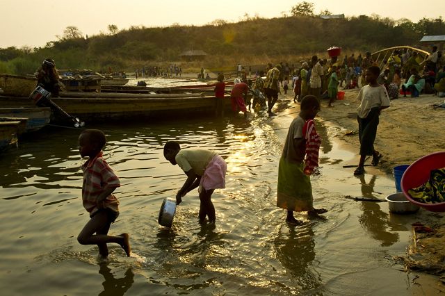 Several villagers stand on a small beach near several small boats; some wade into the water.