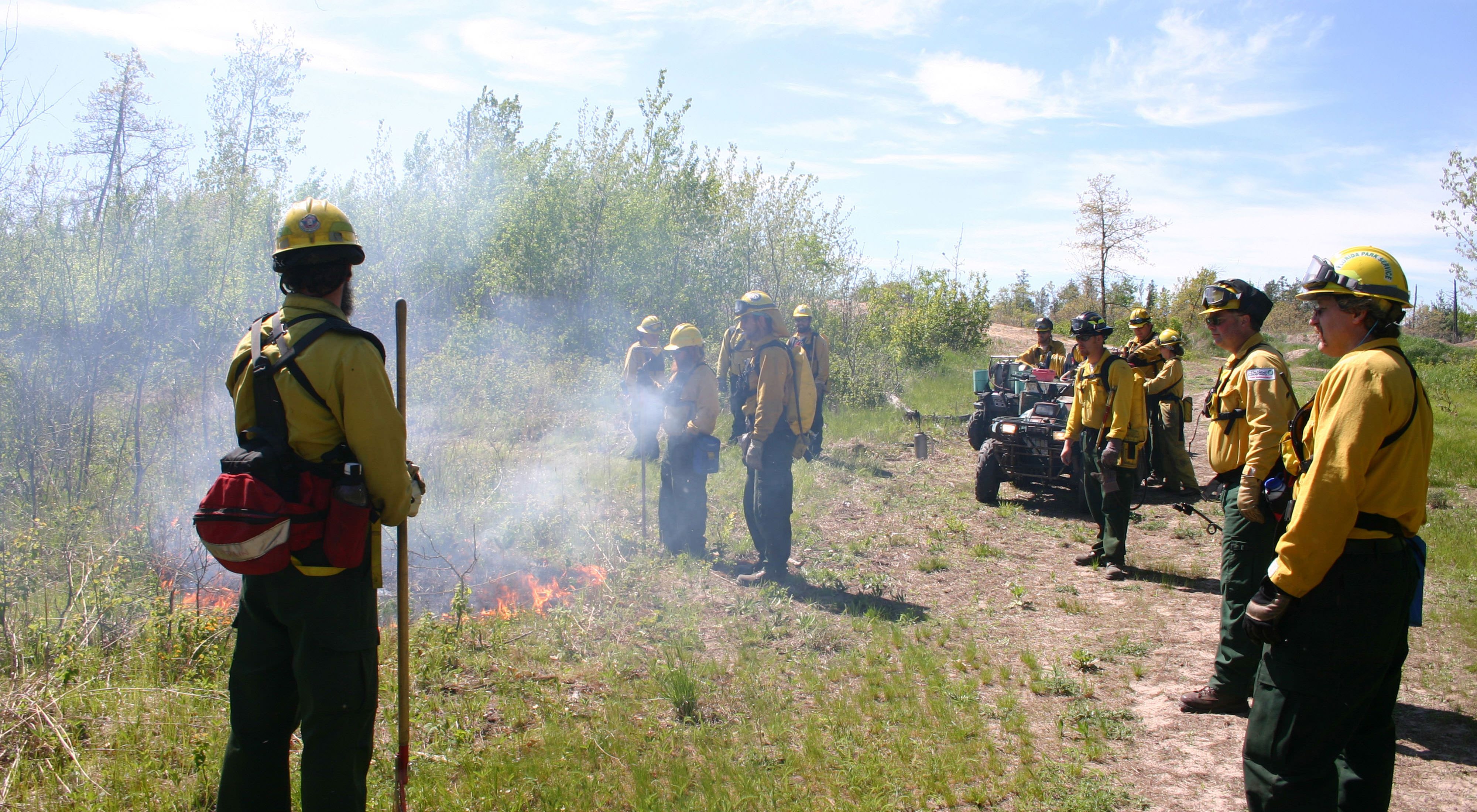 Fire crew members in protective gear stand on a train and observe burning brush.