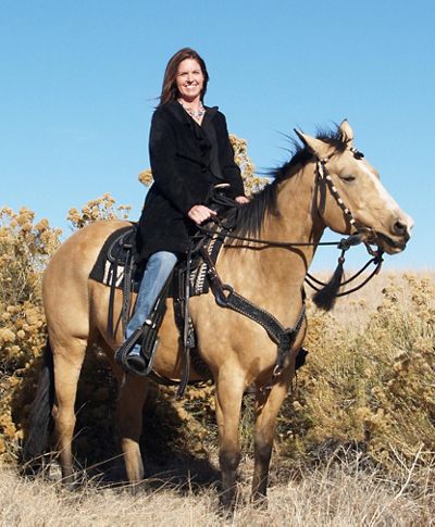 A woman wearing blue jeans and a black jacket sits atop a light brown horse.