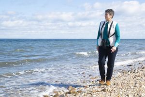 Louise Harrison walking on a shoreline at right looking left to the ocean shore.